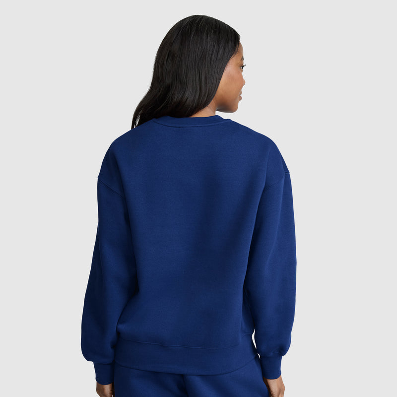A person with long dark hair, seen from behind, stands against a light gray background wearing a plain blue Champion Powerblend Crewneck, Small “C” sweatshirt and matching blue pants.