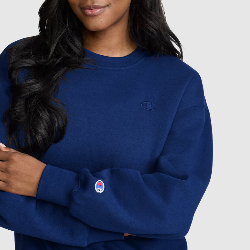 A woman stands against a plain light background with her arms crossed, wearing a dark blue Champion Powerblend Crewneck, Small “C” sweatshirt. This women’s crewneck offers a comfortable fit and features Champion’s signature Powerblend fleece for extra warmth and softness, with distinctive small “C” logo patches on the chest and sleeve.