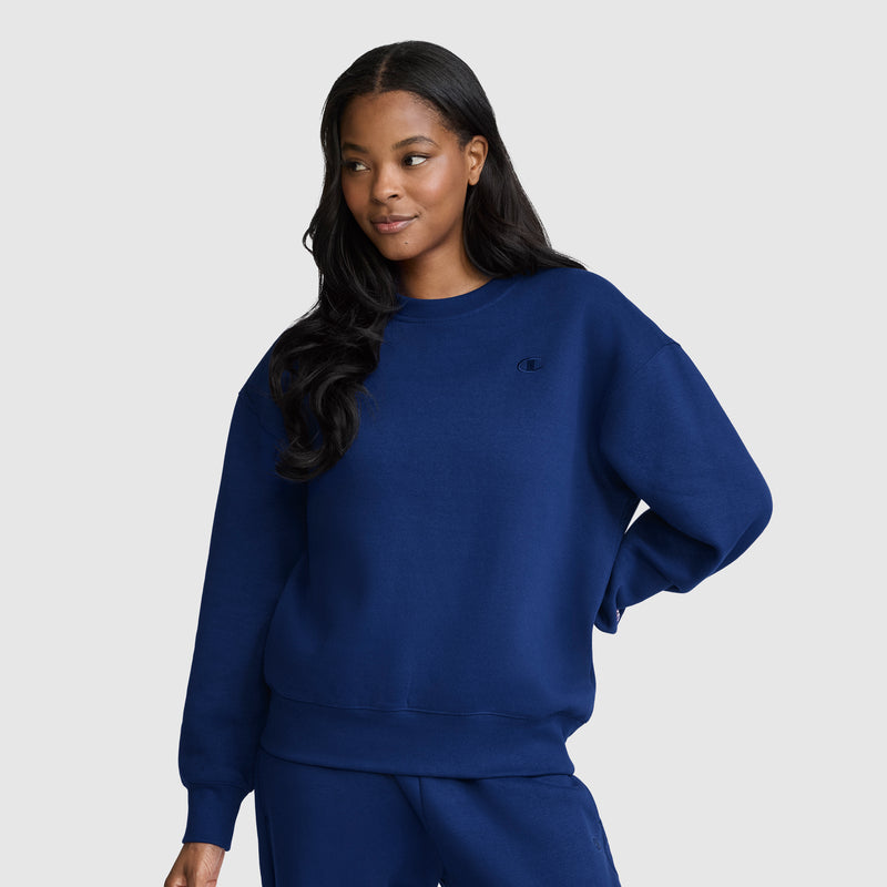 A woman with long dark hair stands against a plain light gray background, wearing a matching navy blue Champion Powerblend Crewneck, Small “C”, and sweatpants. She poses with one hand on her hip and maintains a neutral expression.