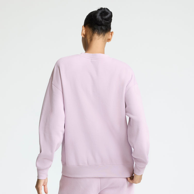 A person with dark hair in a bun is seen from the back, wearing a light pink Champion Powerblend Crewneck with the Small "C" logo and matching fleece pants, showcasing everyday comfort against a plain white background.