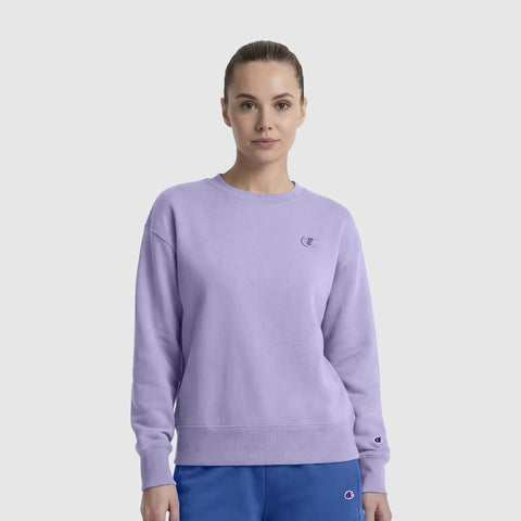 A woman faces forward in a light purple Champion Powerblend Crewneck sweatshirt with a small "C" logo, made from midweight fleece, paired with blue pants. She embodies everyday comfort against a plain white background.