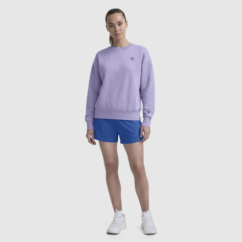 A woman stands facing forward in a light purple Champion Powerblend Crewneck with a small "C" logo, paired with blue shorts and white sneakers, against a plain light gray background.