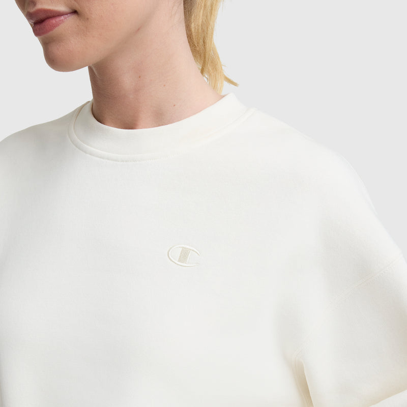 Someone wears a Champion Powerblend Crewneck sweatshirt in off-white, featuring midweight fleece and a small embroidered "C" logo on the chest, shown from the shoulder up against a plain light background.