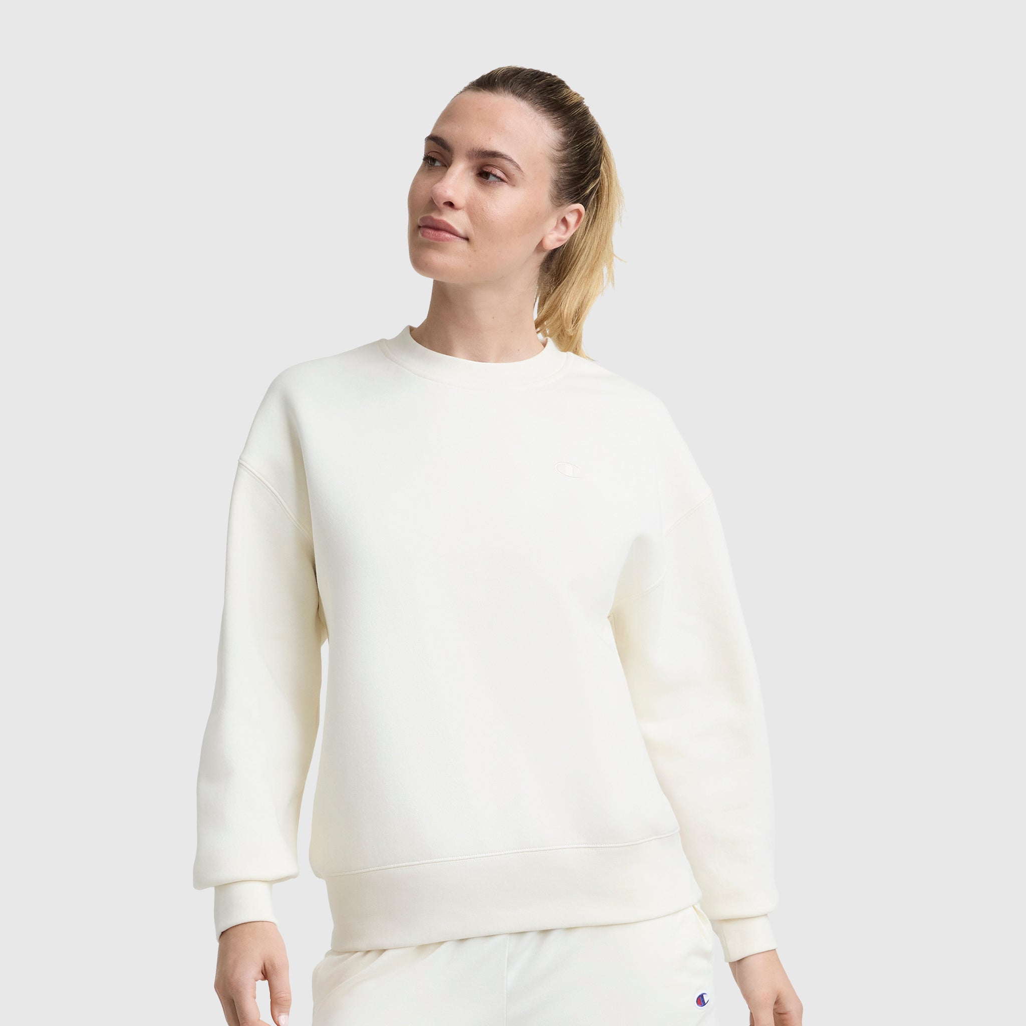 A woman with blonde hair in a ponytail wears the Champion Powerblend Crewneck, Small "C," and matching off-white pants against a plain light gray background. She looks slightly to the side with a neutral expression.