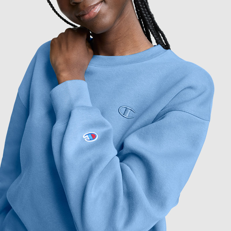 A person wearing a light blue Champion Powerblend Crewneck, Small “C”, touches their collar. Only the lower half of the face is visible, revealing a subtle smile. The plain light gray background highlights the cozy, casual feel of this women’s sweatshirt by Champion, crafted from soft Powerblend fleece.