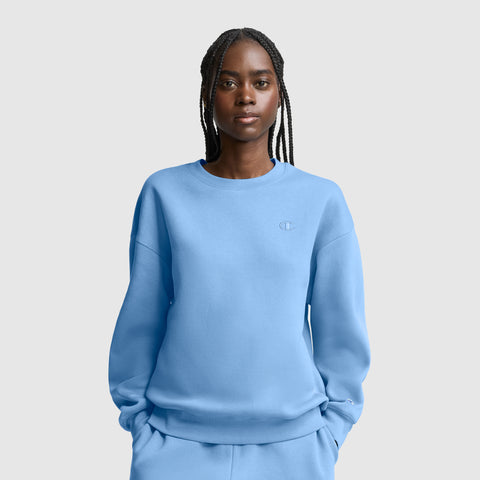 A woman with braided hair wears a light blue Champion Powerblend Crewneck, Small “C”, and matching pants as she stands against a plain light gray background with her hands in her pockets.