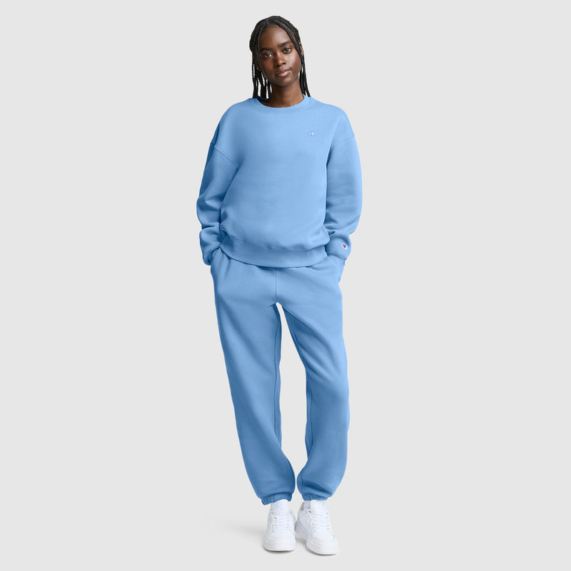 A person stands facing forward, wearing a matching light blue Champion Powerblend Crewneck, Small “C” sweatshirt and sweatpants set with white sneakers, against a plain light gray background.