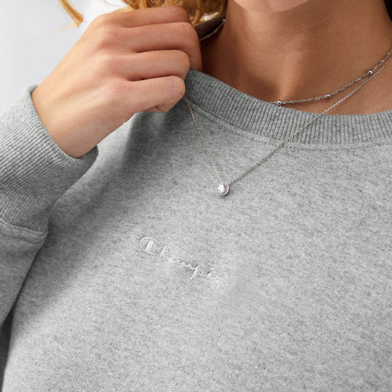 Wearing the Powerblend Crewneck Sweatshirt with Embroidered Script Logo by Champion and two delicate silver necklaces, one with a small pendant, a person lightly touches the collar near their shoulder.