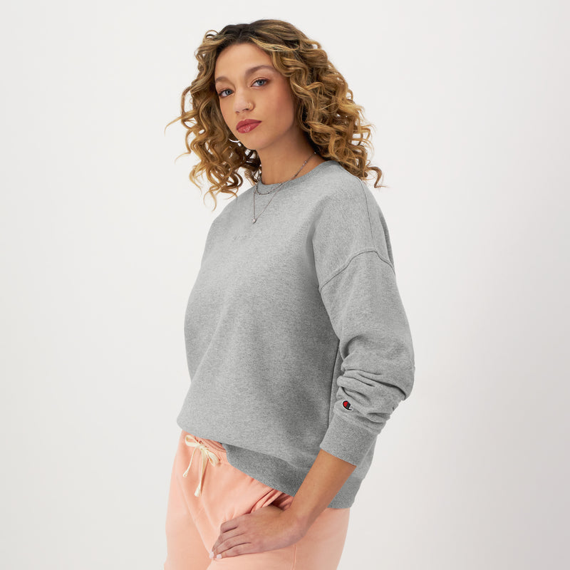 A woman with curly hair wears a Champion Powerblend Crewneck Sweatshirt with an embroidered script logo and light pink pants, posing confidently against a plain white background.