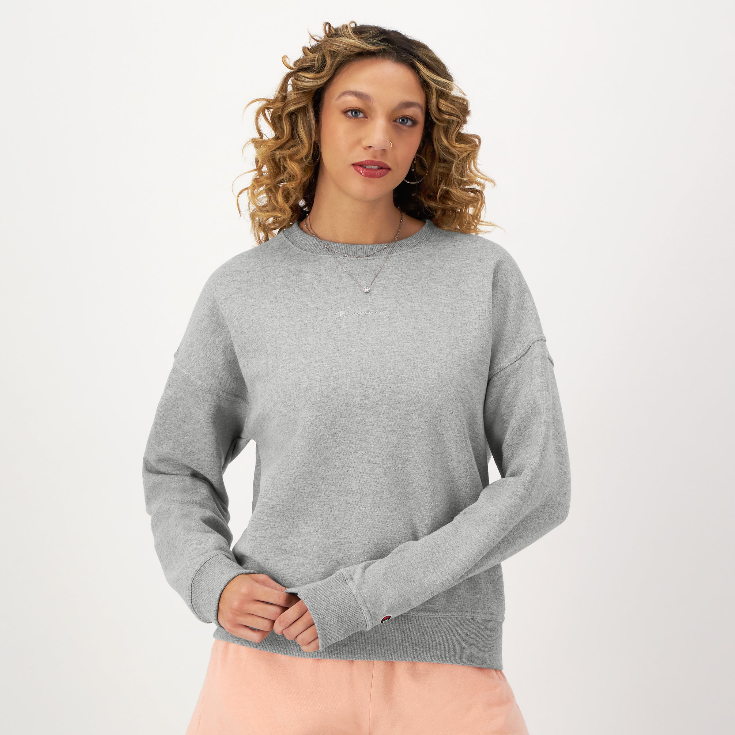 A person with curly hair models the Champion Powerblend Crewneck Sweatshirt with an embroidered script logo and light pink pants, standing against a white background and looking forward with a relaxed expression.