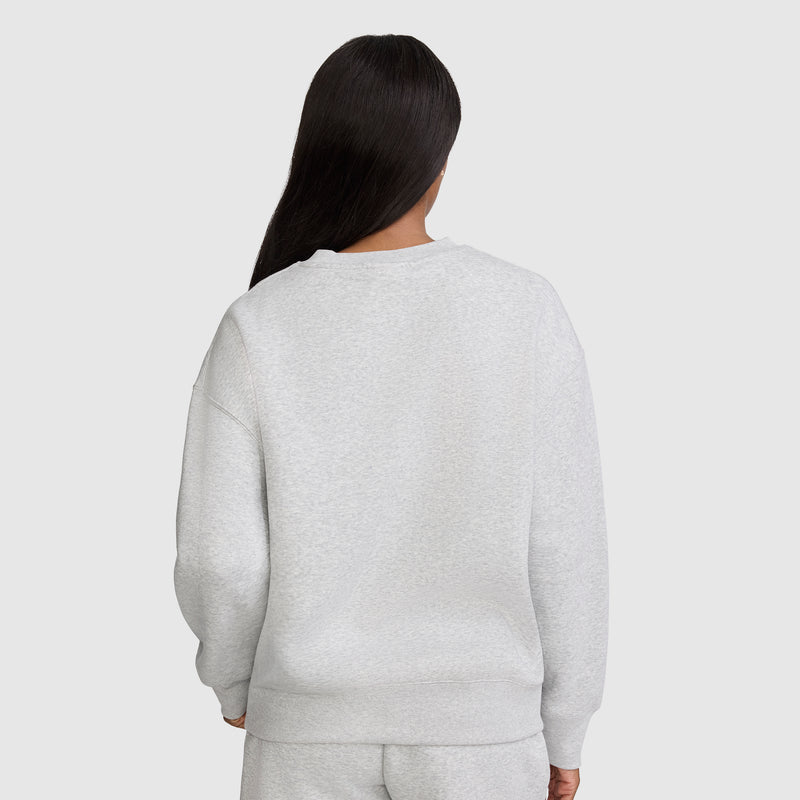 A person with long, straight dark hair is seen from the back, wearing a light gray Champion Powerblend Crewneck Sweatshirt with an embroidered script logo and matching sweatpants against a plain, light gray background, showcasing classic women's fashion in cozy fleece.