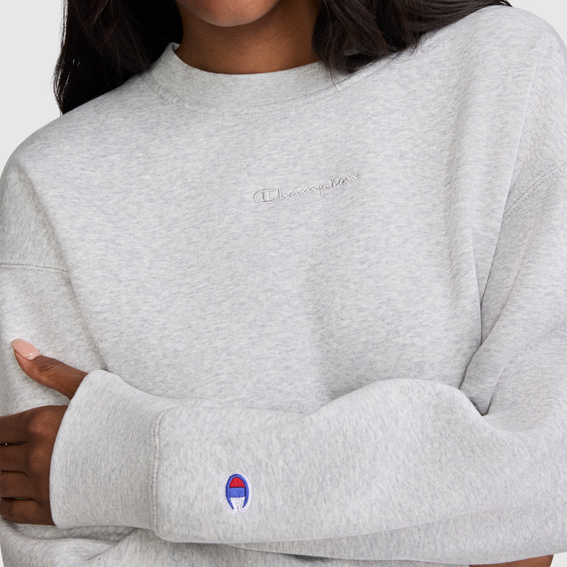 A person wearing a light gray Champion Powerblend Crewneck Sweatshirt with the embroidered script logo on the chest and sleeve. With arms crossed and face partially out of frame, they showcase this cozy staple from Champion's collection of women's crewneck sweatshirts, a popular choice in women's fashion.