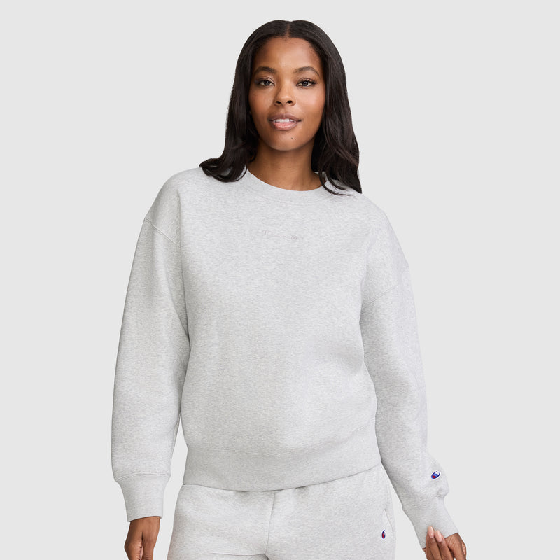 A woman stands against a plain light background, looking at the camera with a slight smile, dressed in a light gray Champion Powerblend Crewneck Sweatshirt with an embroidered script logo and matching sweatpants. This cozy fleece set is an ideal choice for anyone who values comfort and timeless style from Champion's women's collection.