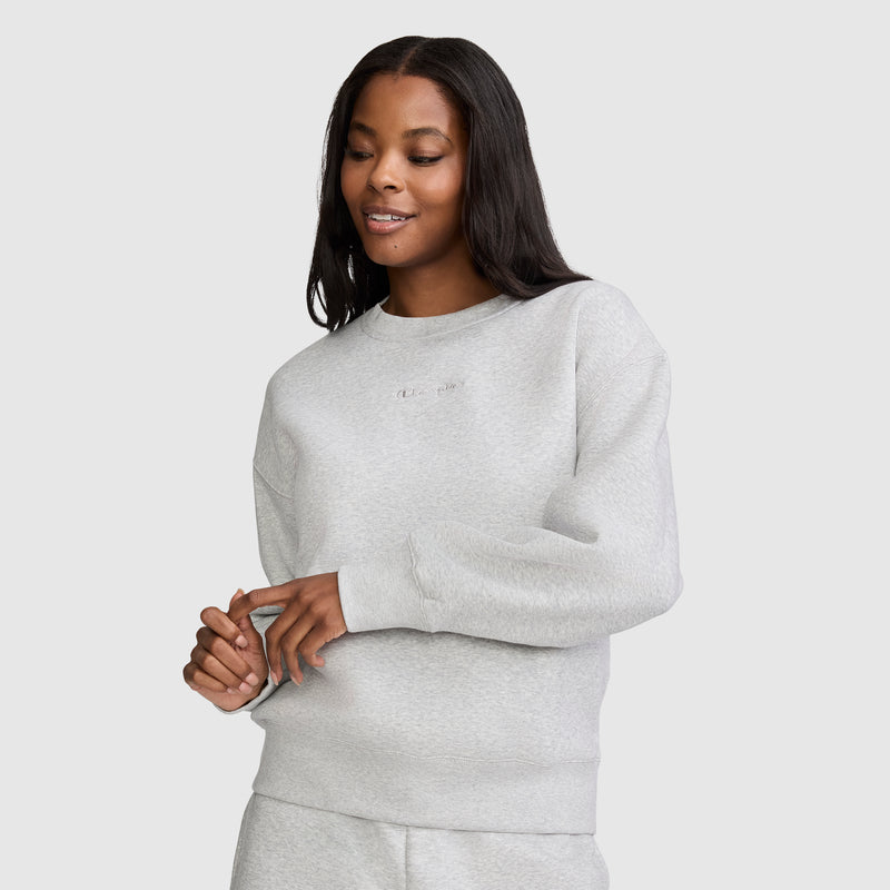A woman with long dark hair wears the Champion Powerblend Crewneck Sweatshirt with Embroidered Script Logo and matching pants, standing against a plain light background and smiling softly with her hands gently clasped in front of her. Crafted for comfort and style, the Powerblend fleece sets this Champion sweatshirt apart in women's fashion.