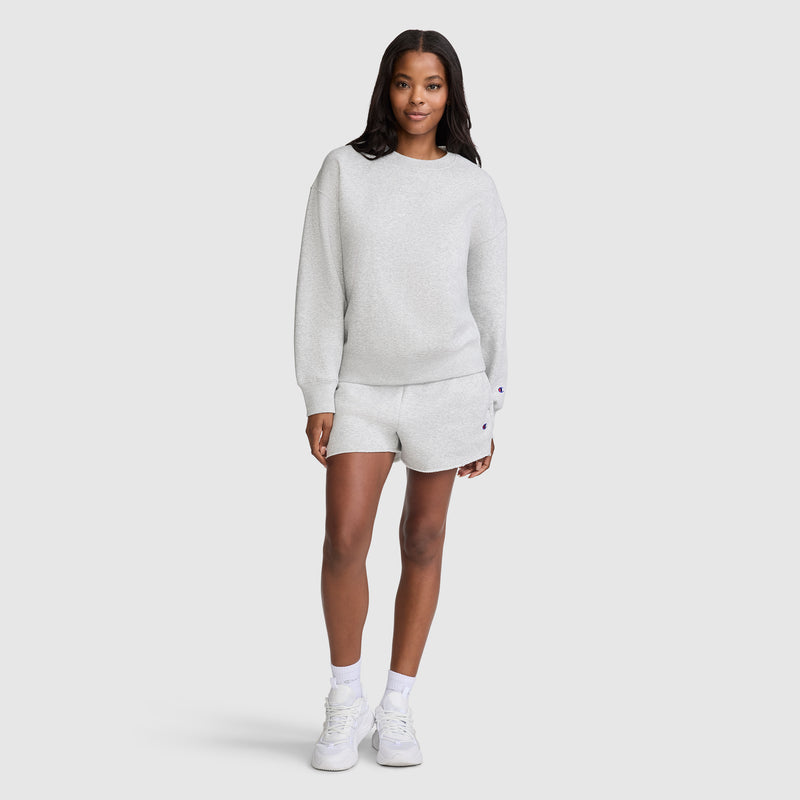 A woman stands facing forward, wearing a light gray Champion Powerblend Crewneck Sweatshirt with an embroidered script logo and matching shorts, both crafted from soft fleece, paired with white sneakers against a plain light background.