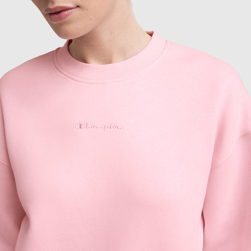 A person wears the Champion Powerblend Crewneck Sweatshirt with an embroidered script logo on the chest. The light pink sweatshirt is shown from the shoulders up against a plain light gray background.