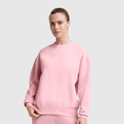 A woman wears the Champion Powerblend Crewneck Sweatshirt, Embroidered Script Logo, in light pink with matching pants against a gray background. The fleece features a chest script logo and a logo patch on the sleeve.