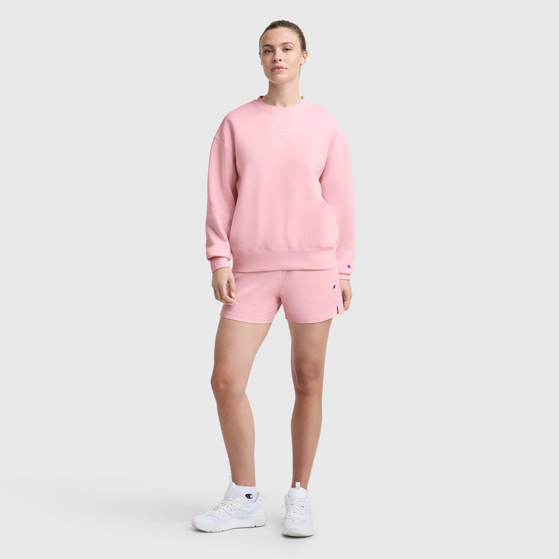 A person stands against a plain gray background, wearing a Champion Powerblend Crewneck Sweatshirt with an embroidered script logo in light pink and matching shorts, paired with white athletic shoes.