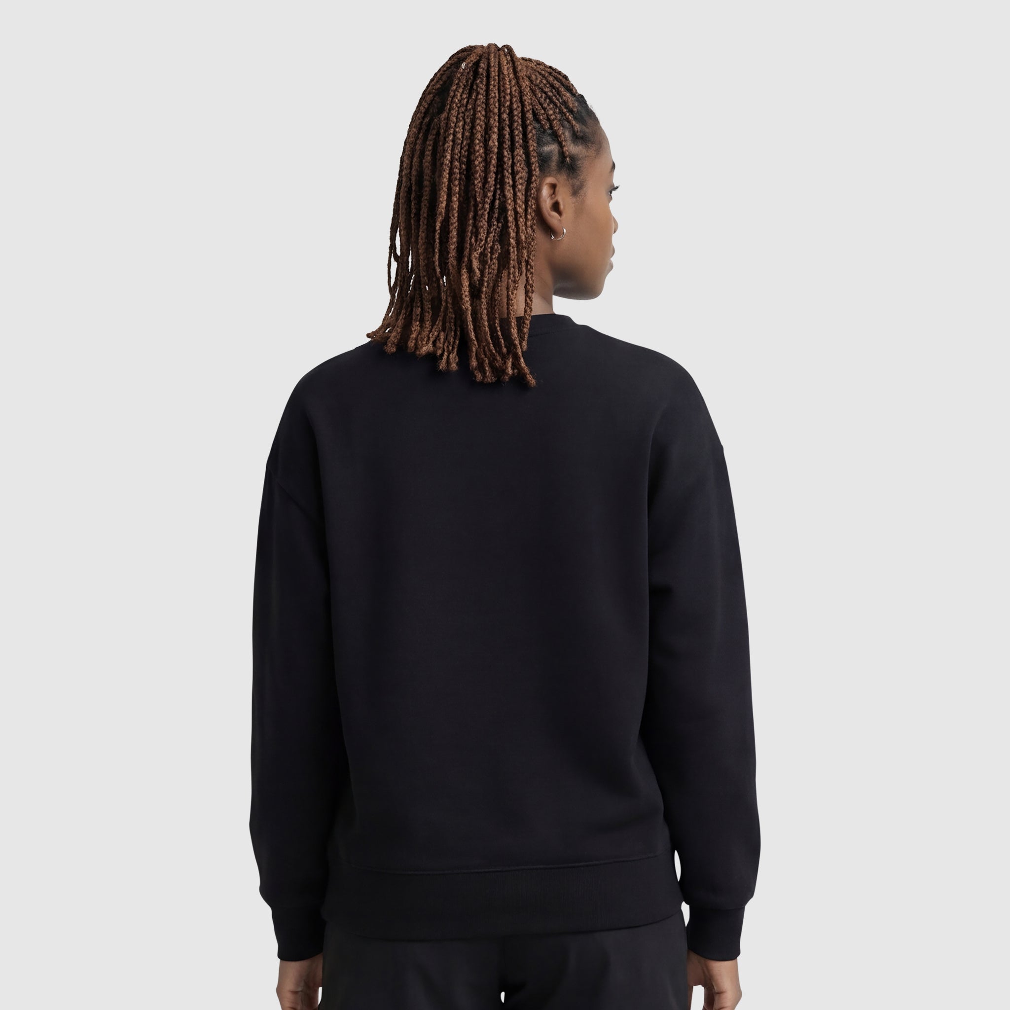 A person with braided hair, seen from behind, is wearing a Champion Powerblend Crewneck Sweatshirt with an embroidered script logo and black pants, standing against a light gray background.