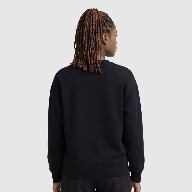 A person with braided hair, seen from behind, is wearing a Champion Powerblend Crewneck Sweatshirt with an embroidered script logo and black pants, standing against a light gray background.