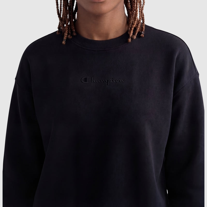 A person with short, braided hair wears a plain black Champion Powerblend Crewneck Sweatshirt with an embroidered script logo on the chest. The background is light gray, and the person's face is partially obscured.