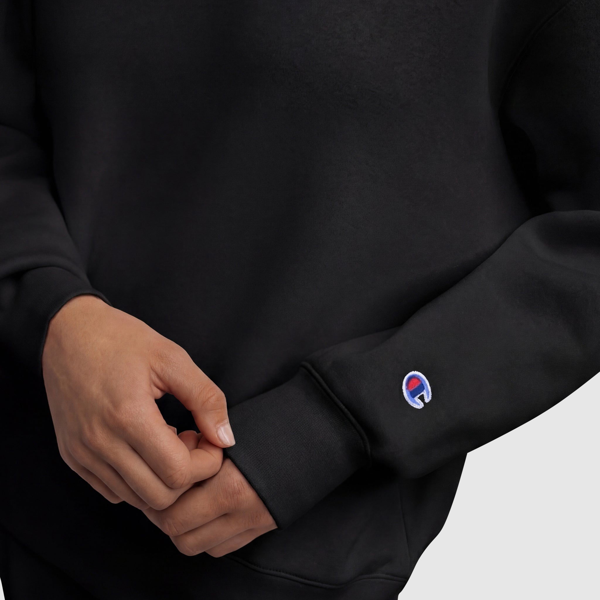 A person in a black Champion Powerblend Crewneck Sweatshirt featuring a small embroidered script logo in blue, white, and red on the sleeve adjusts the cuff amid a plain light gray background.