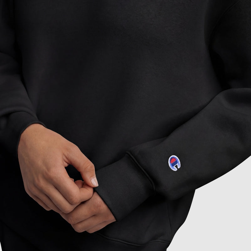 A person in a black Champion Powerblend Crewneck Sweatshirt featuring a small embroidered script logo in blue, white, and red on the sleeve adjusts the cuff amid a plain light gray background.