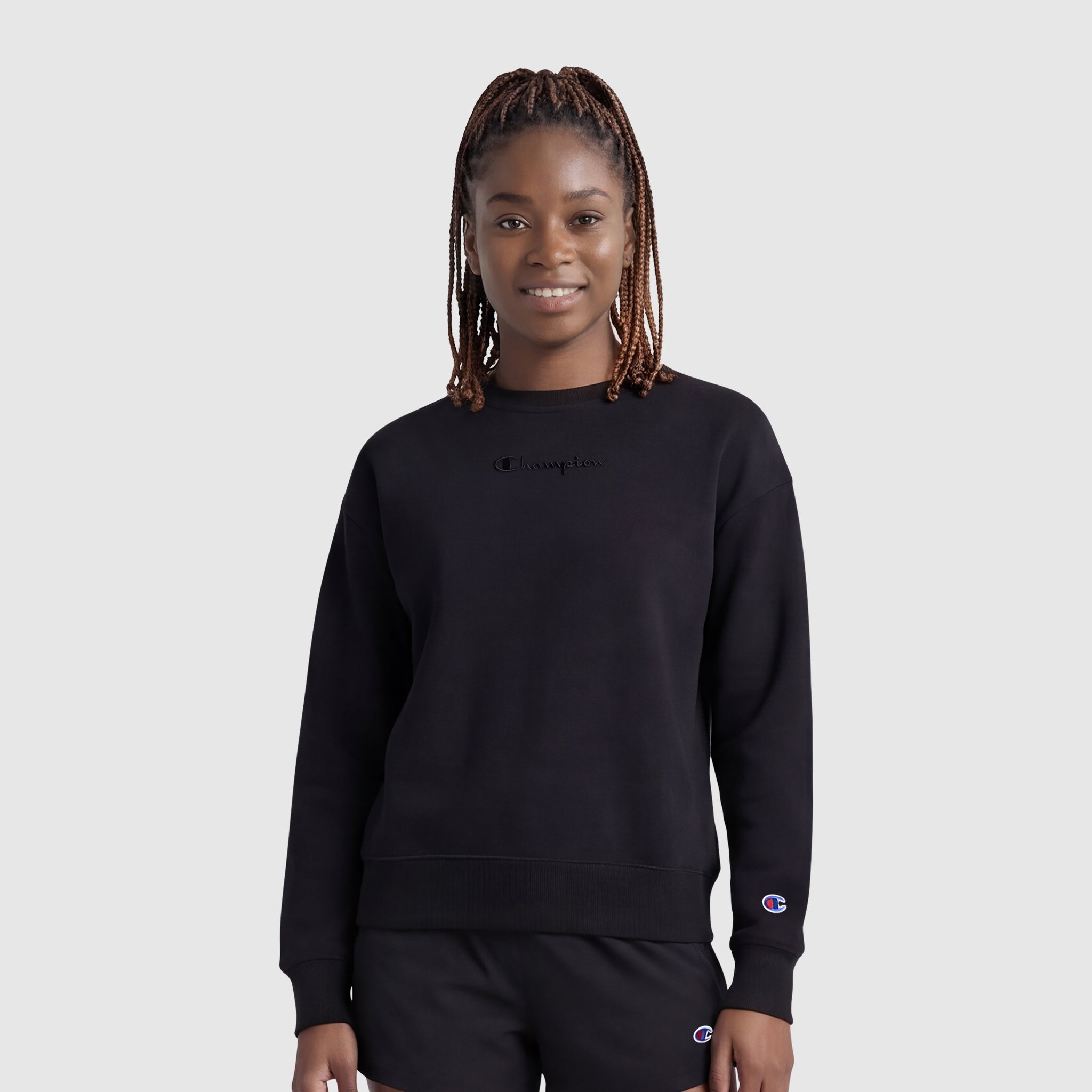 A woman with braided hair smiles while wearing the Champion Powerblend Crewneck Sweatshirt with an embroidered script logo and matching black shorts, standing against a plain light gray background.
