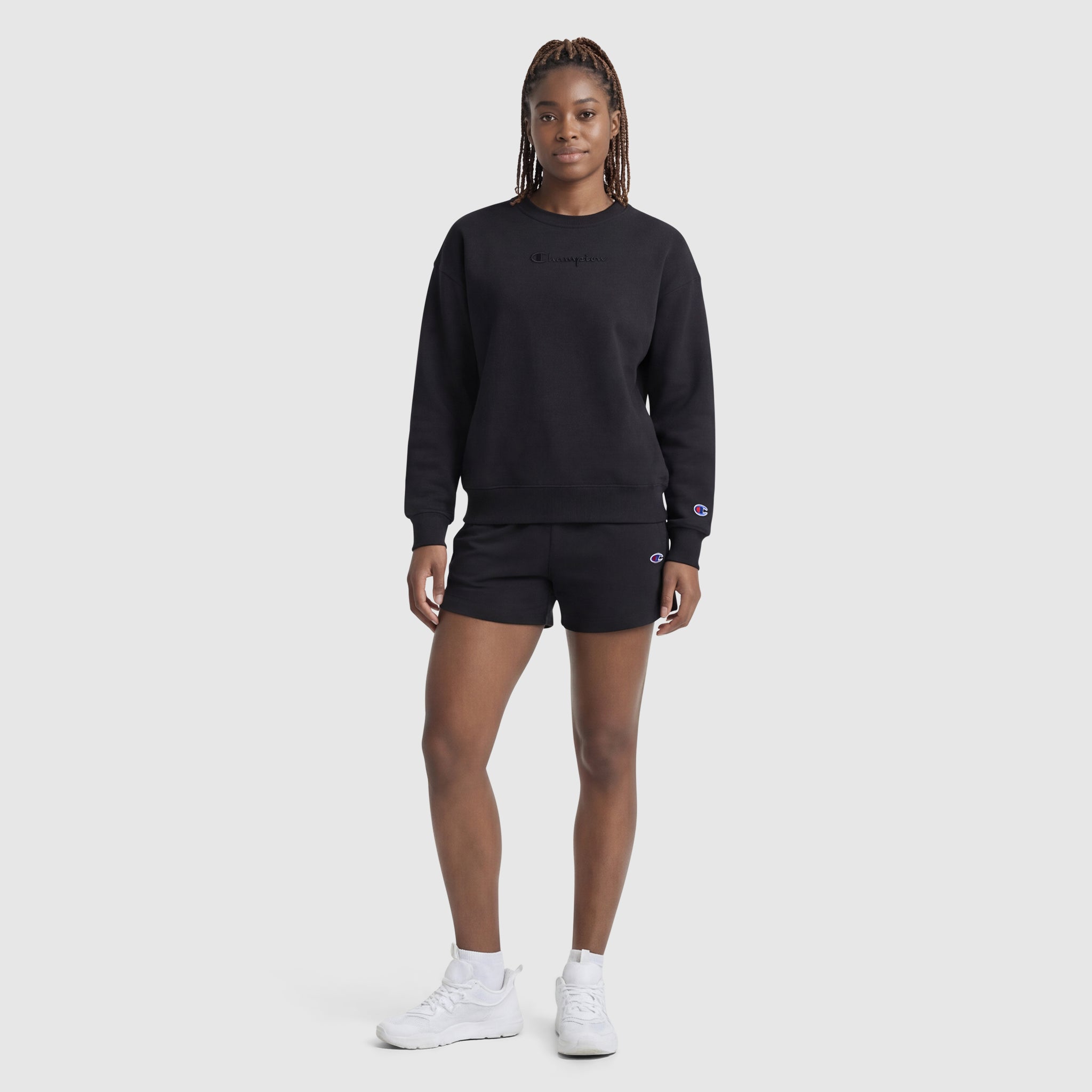 A person with long, braided hair stands against a plain background, wearing a black Champion Powerblend Crewneck Sweatshirt with an embroidered script logo, black shorts, and white sneakers. They face forward with a neutral expression and relaxed posture.