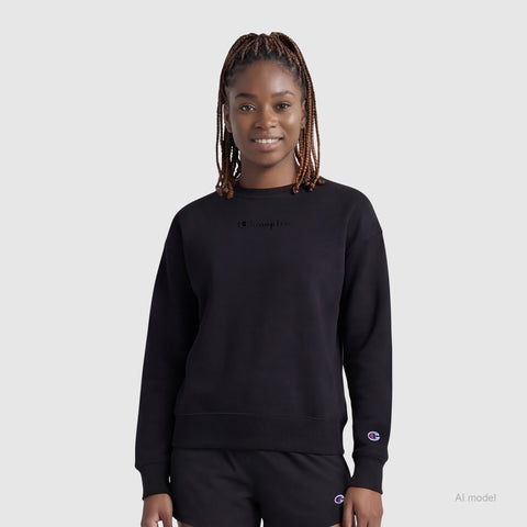 A woman with braided hair wears the Champion Powerblend Crewneck Sweatshirt with embroidered script logo and black shorts, standing against a plain light gray background, facing forward and smiling softly.