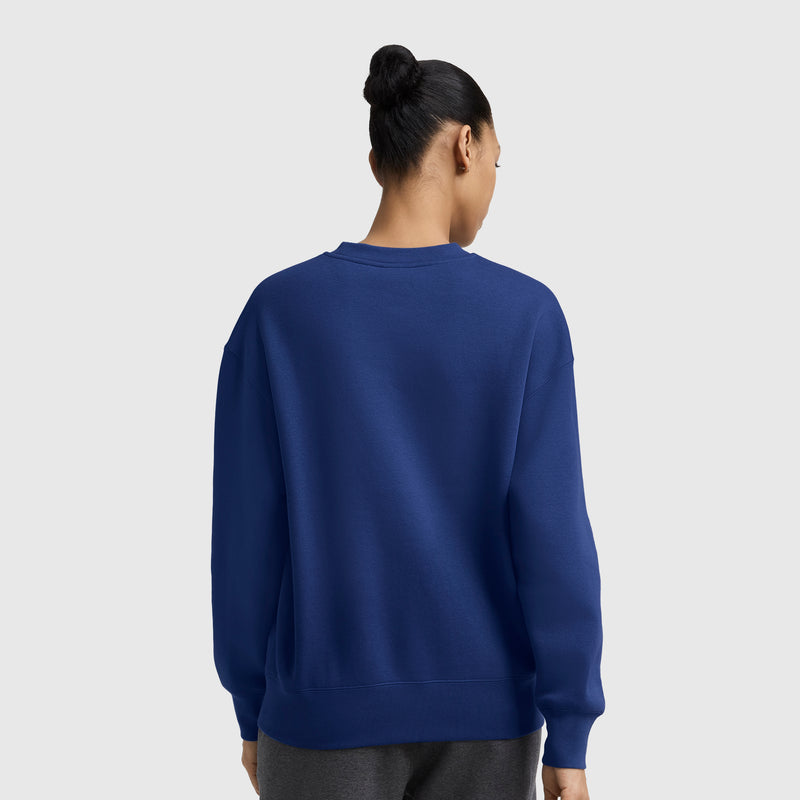 A person with dark hair styled in a bun is seen from the back, wearing a blue Champion Powerblend Crewneck Sweatshirt with an embroidered script logo and dark pants, standing against a plain light gray background.