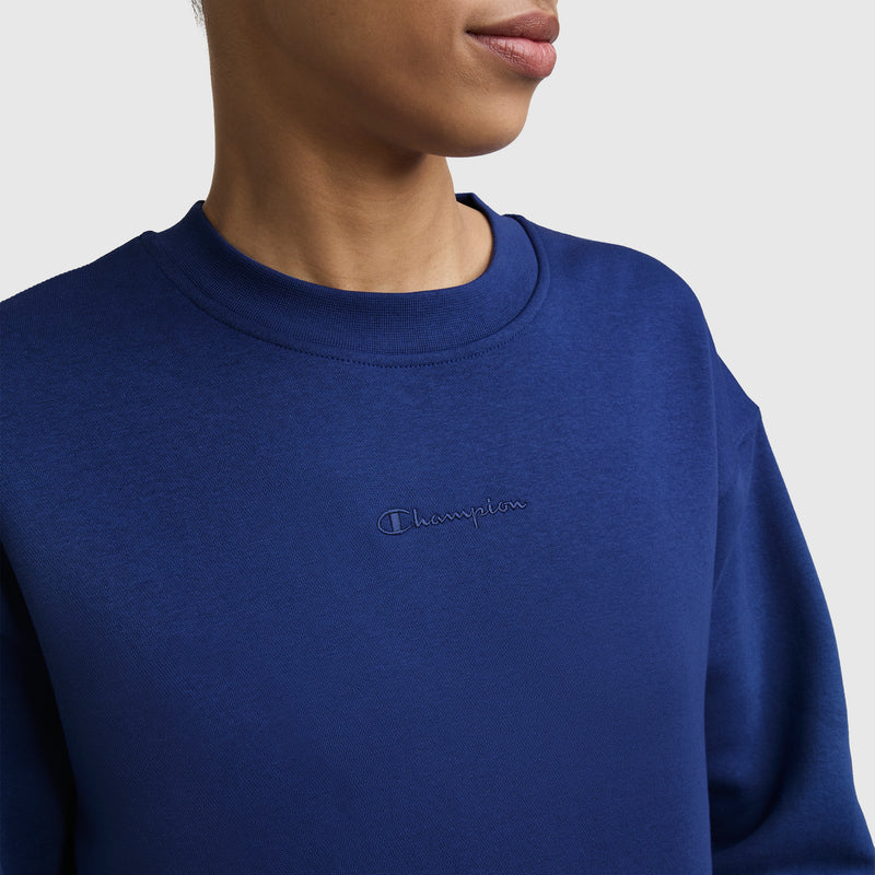 A person is pictured against a simple light backdrop, wearing the Champion Powerblend Crewneck Sweatshirt, Embroidered Script Logo in royal blue. The image focuses on the lower face, neck, and upper torso, showcasing the soft Powerblend fleece fabric and the stylish embroidered script logo on the chest.