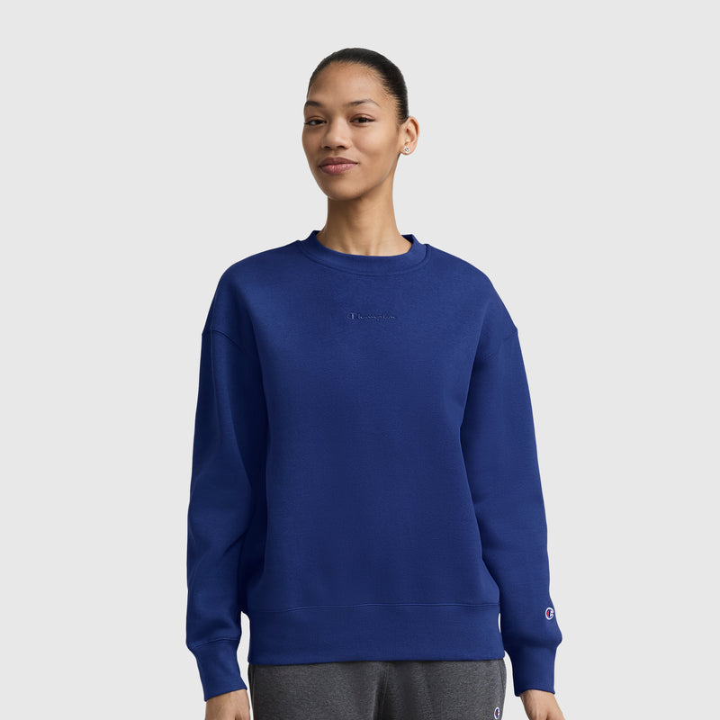 A person stands against a plain white background, smiling slightly with hands relaxed at their sides, wearing a Champion Powerblend Crewneck Sweatshirt with an embroidered script logo in blue and gray pants.