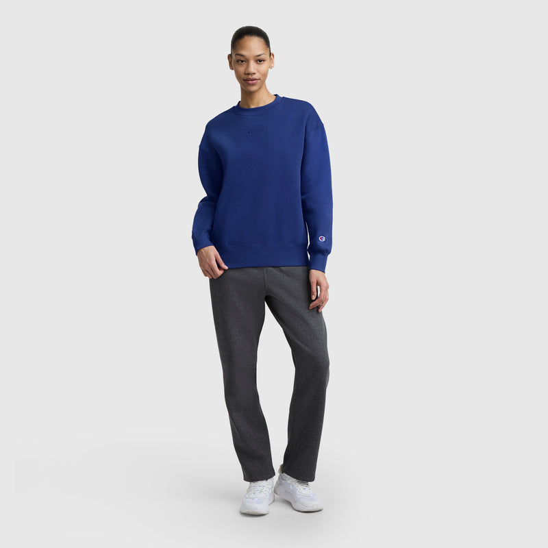 A person stands against a light gray background wearing a Champion Powerblend Crewneck Sweatshirt with an embroidered script logo in blue, paired with gray pants and white sneakers, one hand in their pocket and a slight smile on their face.