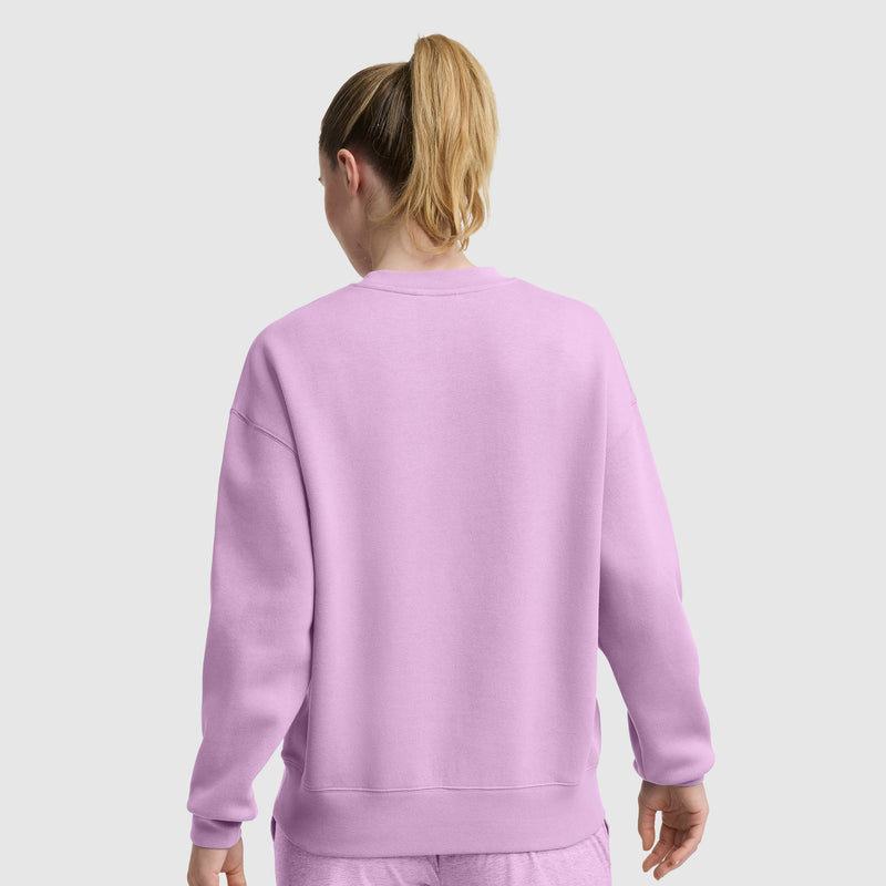 A person with blonde hair in a ponytail is wearing a light purple Champion Powerblend Crewneck Sweatshirt with an embroidered script logo and matching pants, facing away from the camera against a plain light background.
