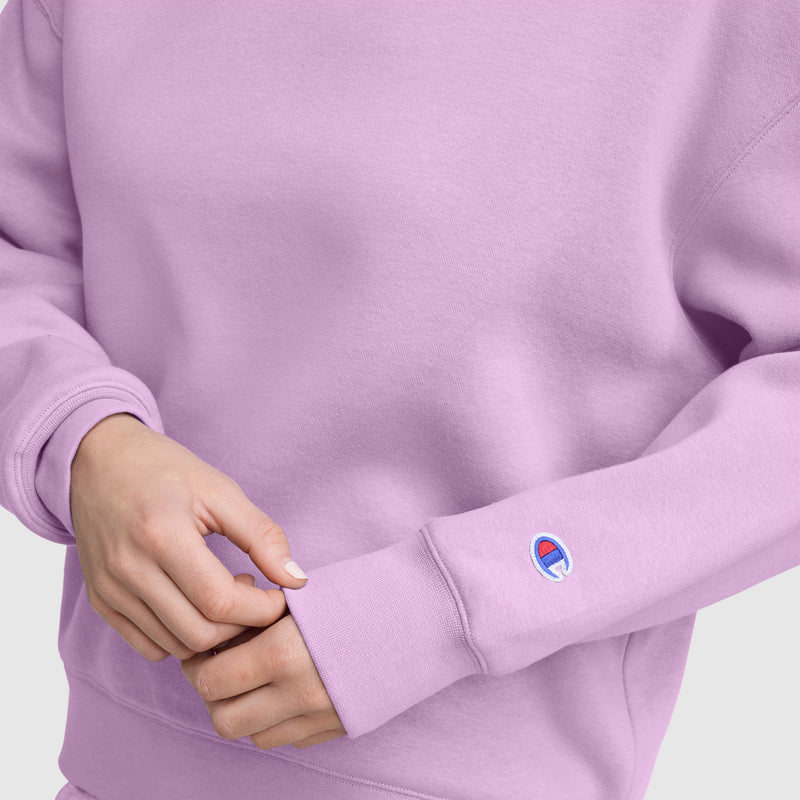 A person wearing a light purple Champion Powerblend Crewneck Sweatshirt, featuring the embroidered script logo on the sleeve, adjusts the cuff with their left hand. The cozy Powerblend fleece provides comfort, set against a plain, neutral background.