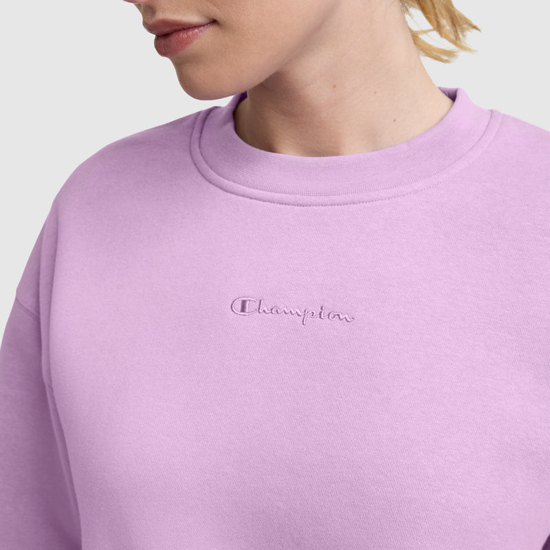 A person is shown wearing a light purple Champion Powerblend Crewneck Sweatshirt with the Embroidered Script Logo. Only their lower face, neck, and upper torso are visible. The cozy fleece sweatshirt features the Champion logo embroidered on the front.