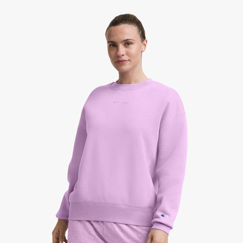 A woman wearing a light purple Champion Powerblend Crewneck Sweatshirt with an embroidered script logo and matching pants stands against a plain white background, looking forward with a slight smile.