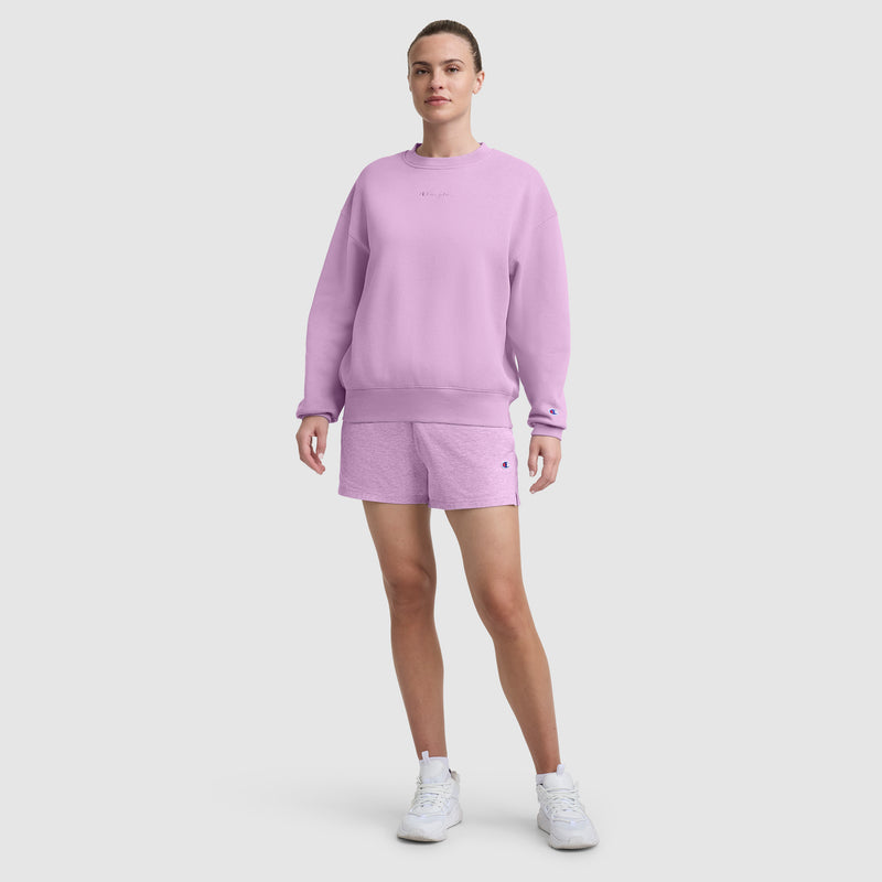 A person stands facing forward, wearing a light purple Champion Powerblend Crewneck Sweatshirt with Embroidered Script Logo and matching shorts, paired with white sneakers, against a plain white background.