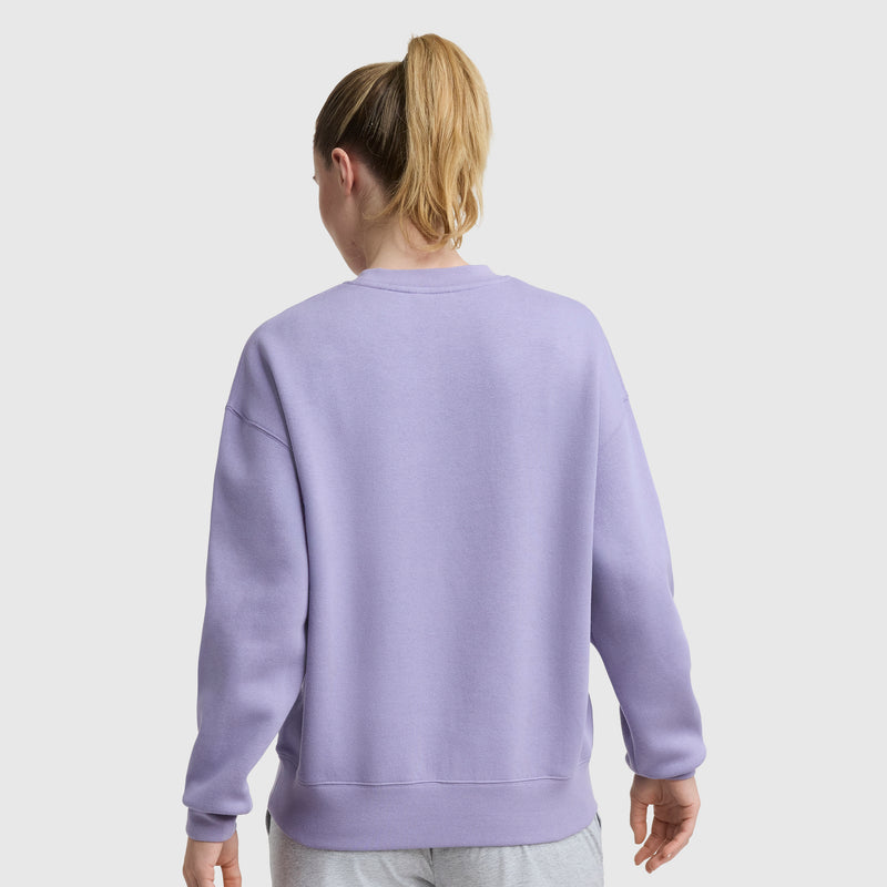 A woman with blonde hair in a ponytail is seen from the back, wearing a light purple Champion Powerblend Crewneck Sweatshirt with an embroidered script logo and light gray pants, standing against a plain white background.