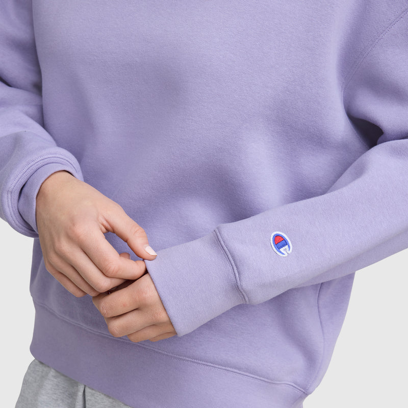 A person in a light purple Champion Powerblend Crewneck Sweatshirt, featuring an embroidered script logo on the sleeve, adjusts the cuff with one hand against a plain light background.