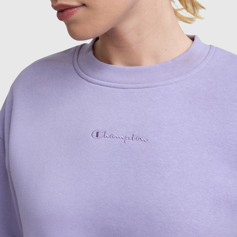 A person wears the Champion Powerblend Crewneck Sweatshirt in light purple, featuring an embroidered script logo at the center chest, against a plain white background.
