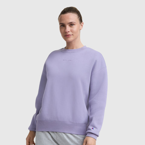 A woman with brown hair in a bun wears the Champion Powerblend Crewneck Sweatshirt, Embroidered Script Logo, in light purple with gray pants, standing against a plain white background. The fleece features a small embroidered logo on chest and sleeve.