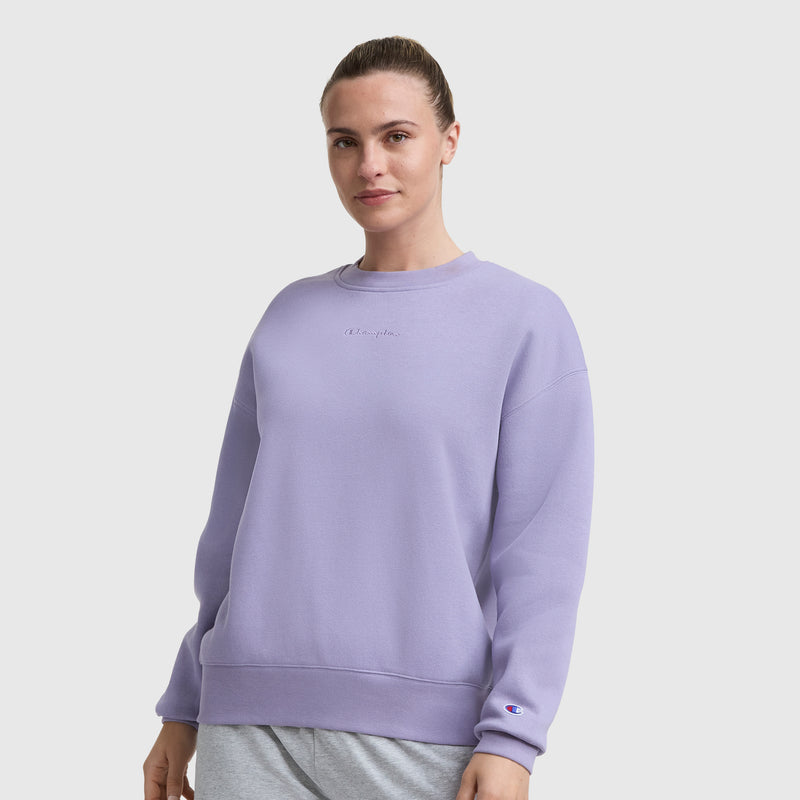 A woman with brown hair in a bun wears the Champion Powerblend Crewneck Sweatshirt, Embroidered Script Logo, in light purple with gray pants, standing against a plain white background. The fleece features a small embroidered logo on chest and sleeve.