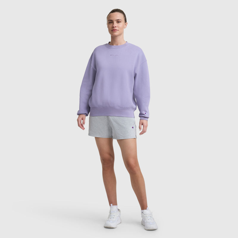 A person stands against a white background wearing the Champion Powerblend Crewneck Sweatshirt with an embroidered script logo in lavender, paired with light gray shorts and white sneakers. Their short hair is pulled back as they look forward neutrally.