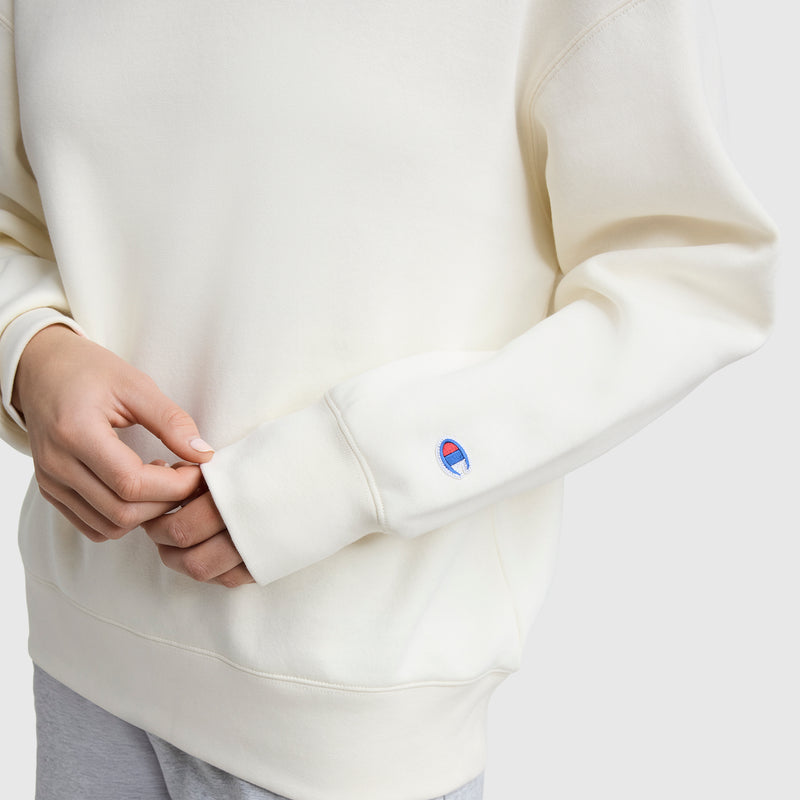 A person in a cream Powerblend Crewneck Sweatshirt by Champion, featuring an embroidered script logo near the wrist, stands against a plain background wearing light gray pants and gently holds the sleeve.