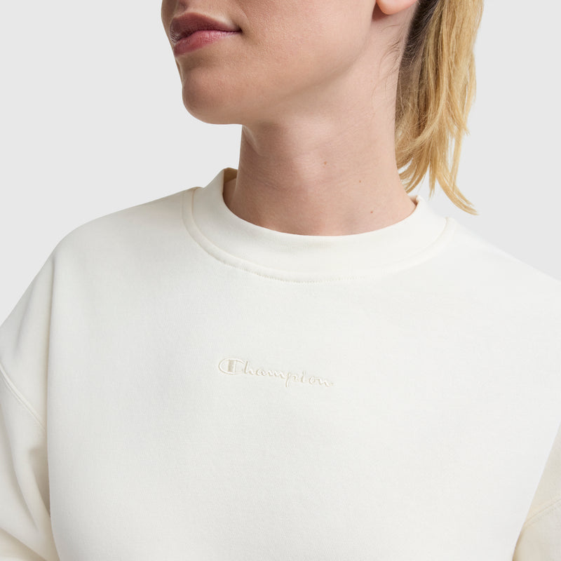 A light-skinned, blonde-haired person wears a loose-fit Champion Powerblend Crewneck Sweatshirt with an embroidered script logo, shown from upper torso to lower face against a plain light background.