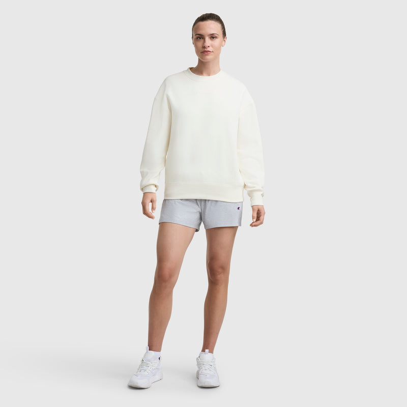 A person stands facing forward wearing a loose-fit white Champion Powerblend Crewneck Sweatshirt with Embroidered Script Logo, light gray shorts, and white sneakers against a plain light background.