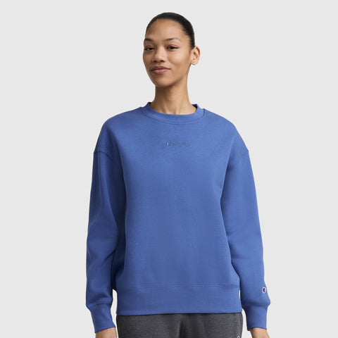 A woman wears the Champion Powerblend Crewneck Sweatshirt with an embroidered script logo and dark gray pants, standing against a light gray background and smiling slightly.