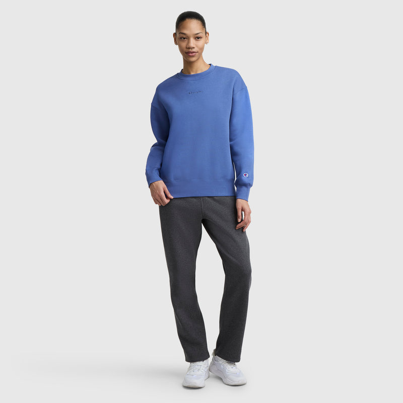 A person stands facing forward in a blue Champion Powerblend Crewneck Sweatshirt with an embroidered script logo, dark gray pants, and white sneakers, against a plain light gray background.